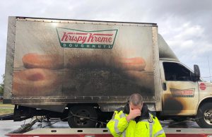 a burnt krispy kreme truck on a truck bed and a police officer in front holding his face