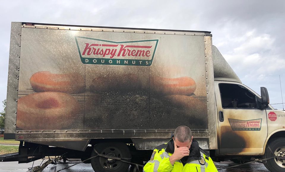a burnt krispy kreme truck on a truck bed and a police officer in front holding his face