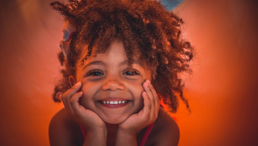 Little Ones Fundraiser: little girl smiling with a little afro and a burnt orange background