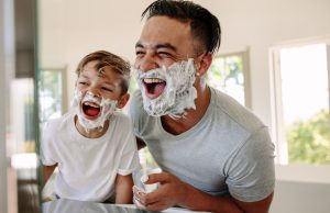Man and little boy with shaving foam on their faces looking into the bathroom mirror and laughing. Father and son having fun while shaving in bathroom.